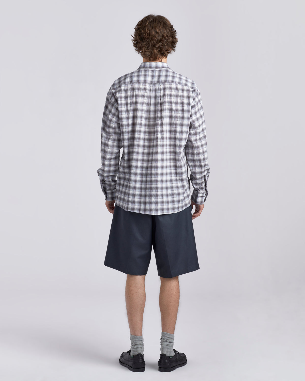 A model shown from the back wearing a gray and white plaid button-down shirt, navy blue shorts, gray socks, and black shoes, photographed against a light gray studio background.