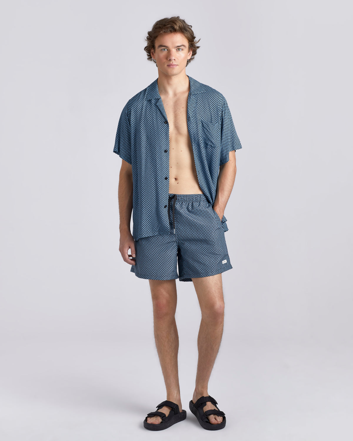 A man modeling a matching two-piece lounge set consisting of a short-sleeve button-up shirt and shorts in a blue and white micro-pattern, paired with black slide sandals, against a light gray studio background.