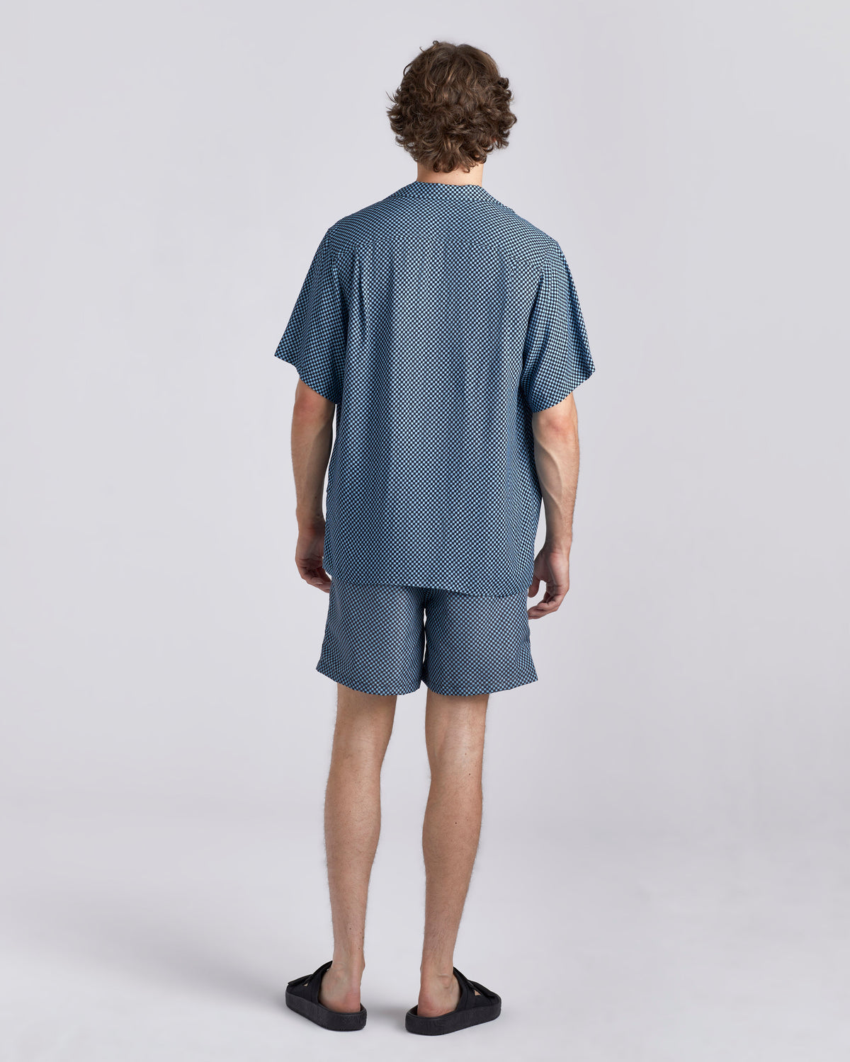 A man stands with his back to the camera wearing a matching blue patterned short-sleeve shirt and shorts set, paired with black slip-on shoes, against a light gray studio background.
