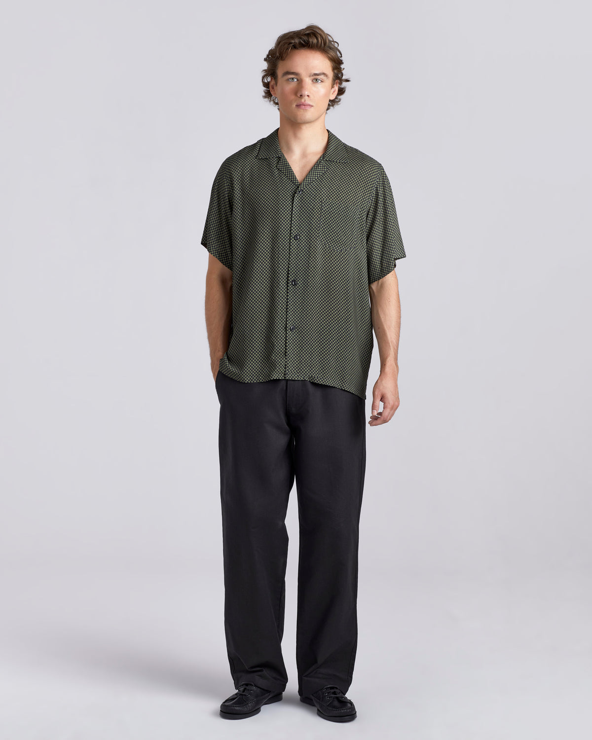 A man modeling a short-sleeve button-up shirt with a fine green and black micro-pattern, paired with black trousers and black shoes, against a light gray studio background.