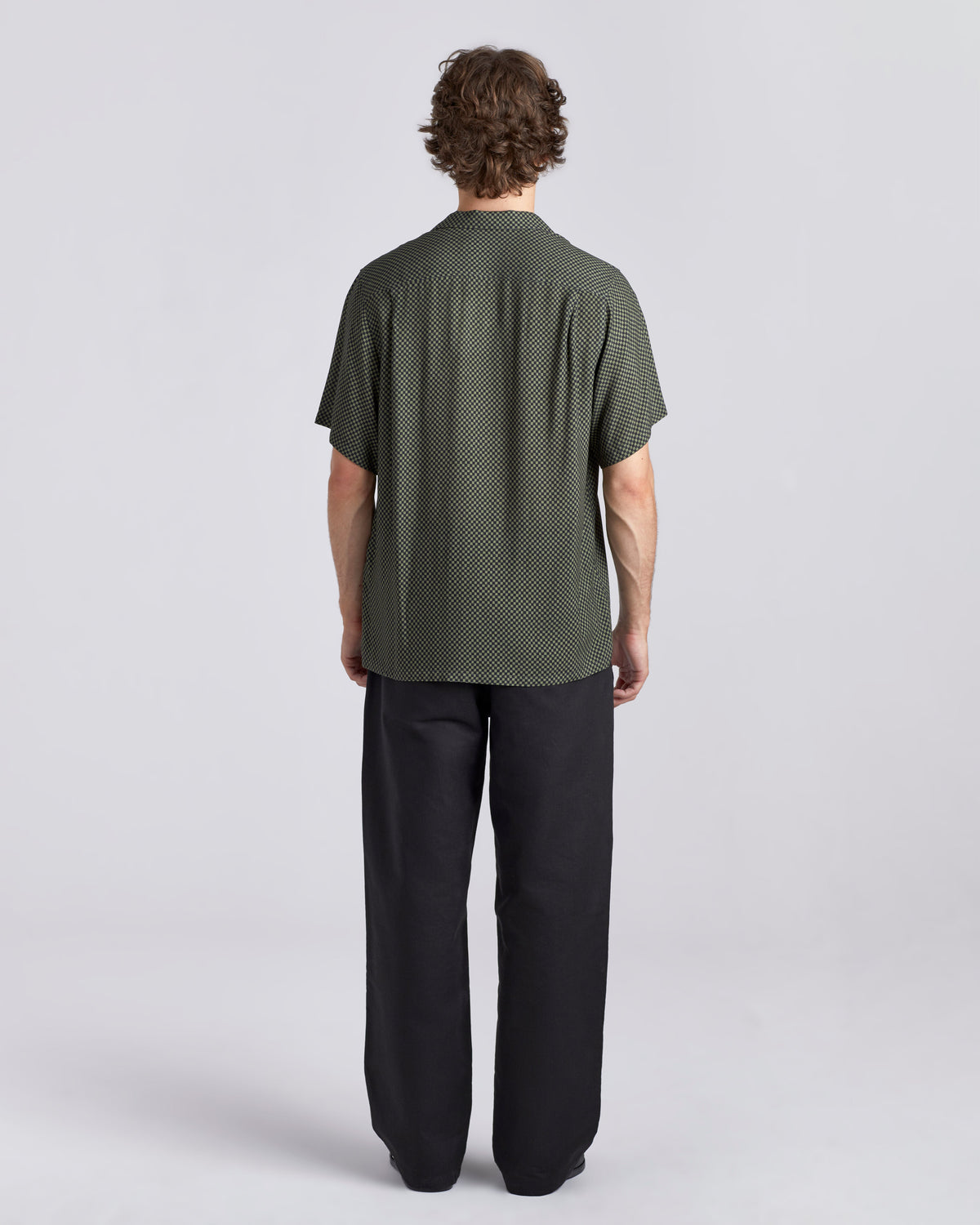 A man viewed from the back wearing a short-sleeve dark green shirt with a subtle textured pattern and black trousers, standing against a light gray studio background.
