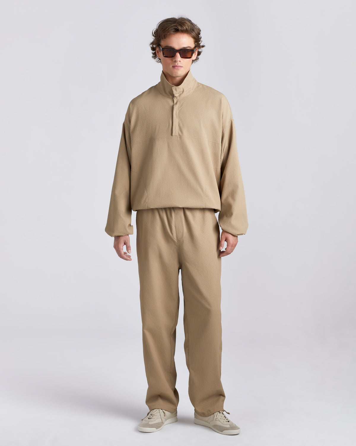 A model wearing a matching beige two-piece outfit consisting of a long-sleeve half-placket pullover and relaxed-fit trousers, styled with beige sneakers and dark sunglasses against a light gray studio background.