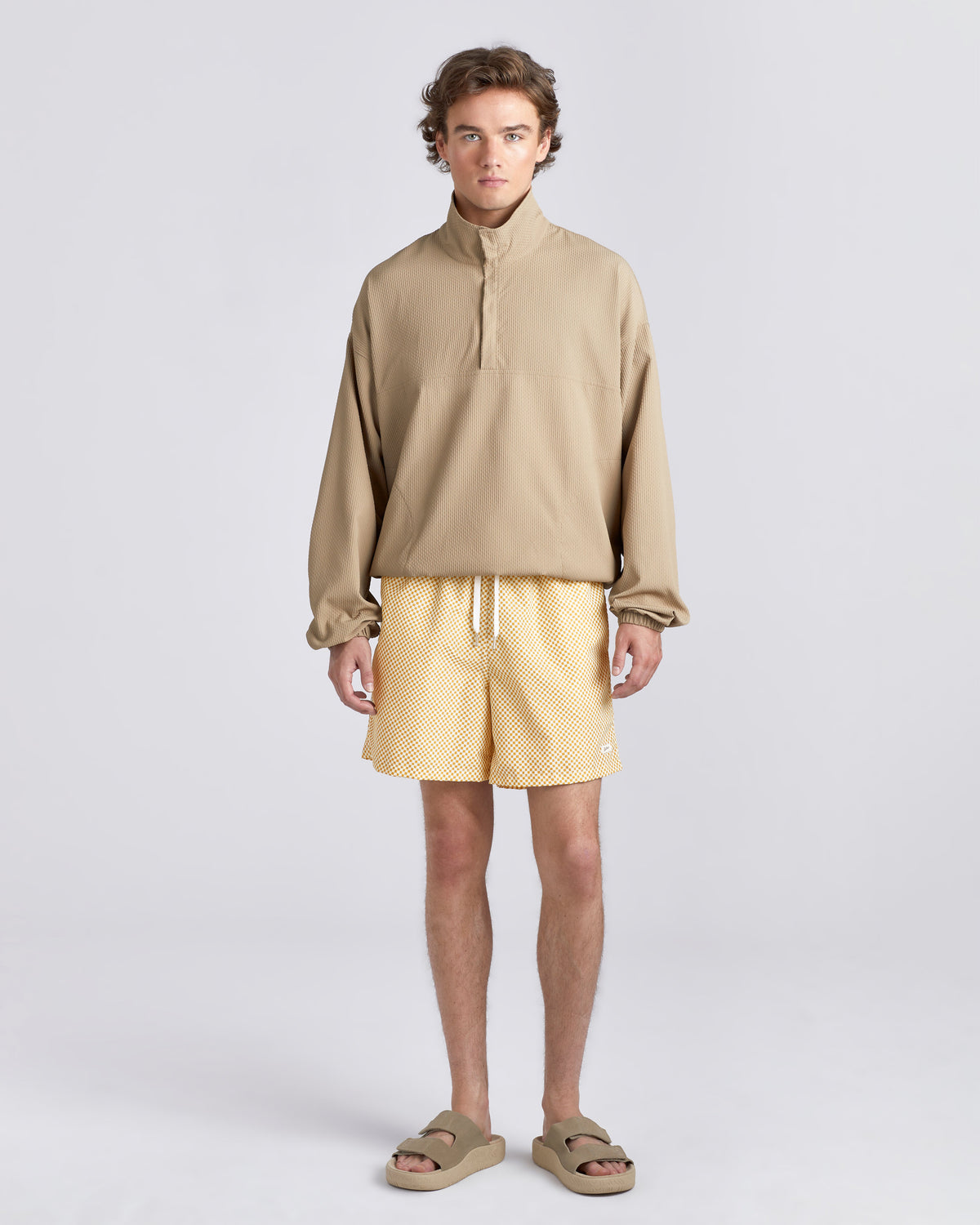 A male model stands against a light gray studio background wearing a beige long-sleeve quarter-zip pullover, light yellow textured shorts, and beige slide sandals.