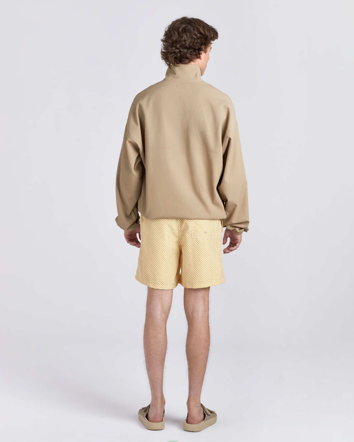 A man shown from the back wearing a beige textured long-sleeve quarter-zip pullover, light yellow textured shorts, and beige slide sandals, set against a plain light gray studio background.