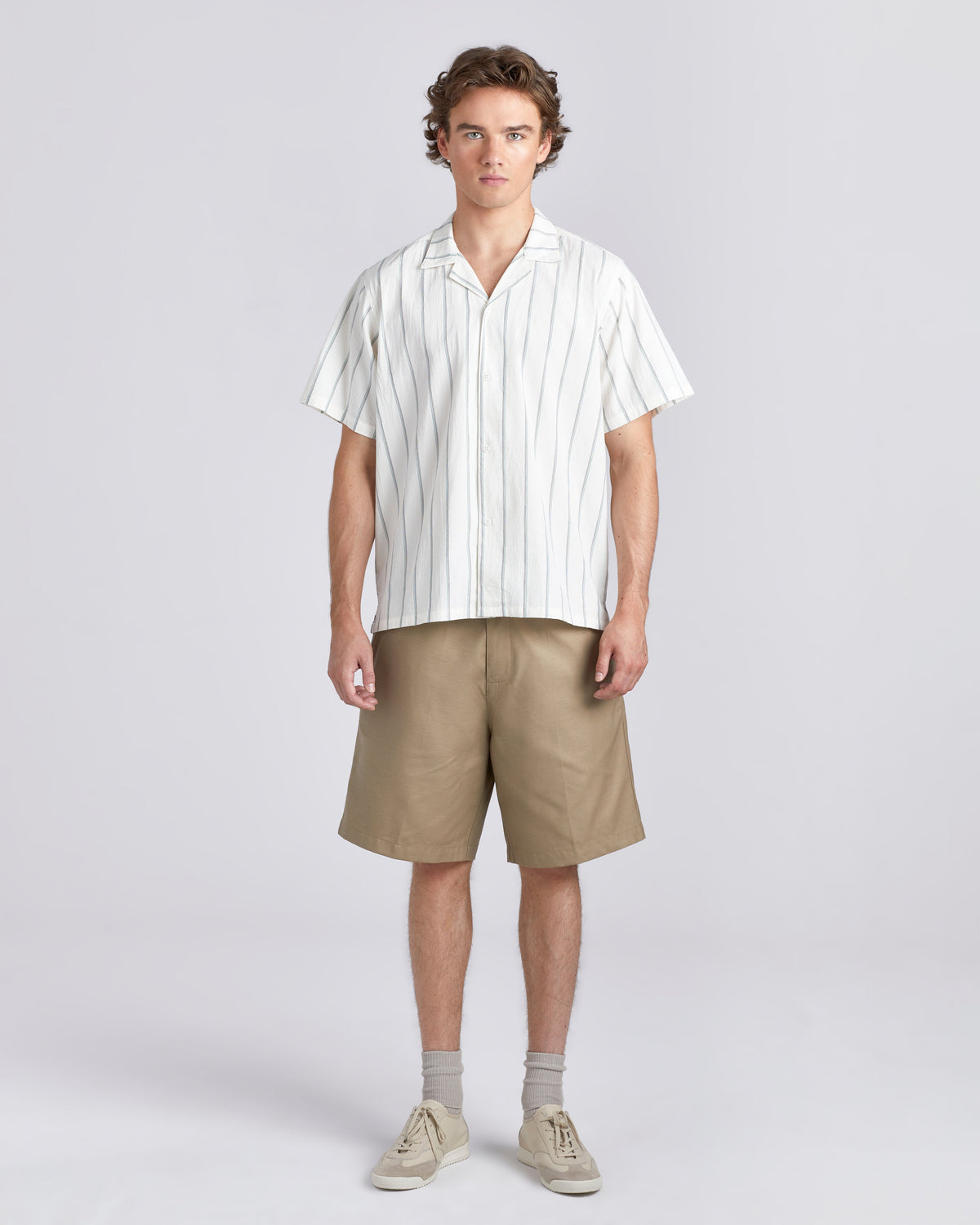 A man stands against a light gray studio background wearing a white short-sleeve button-up shirt with thin vertical tan stripes, khaki shorts, light gray socks, and beige sneakers.