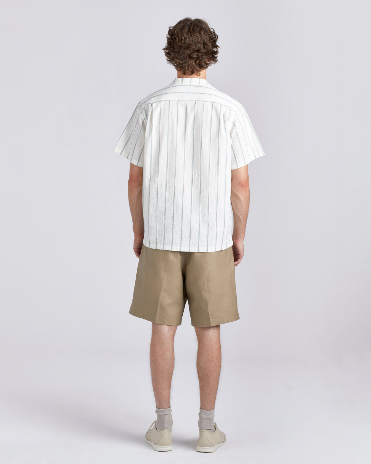 A man viewed from the back wearing a white short-sleeve button-up shirt with thin vertical tan stripes, khaki shorts, and light-colored sneakers against a light gray studio background.