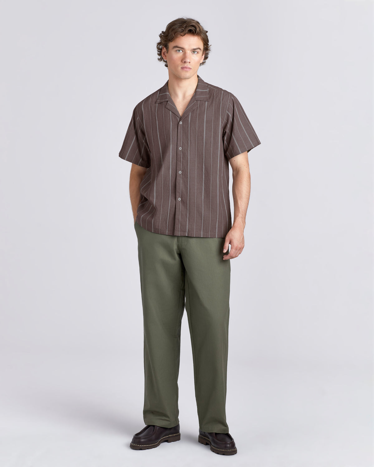 A man modeling a short-sleeve button-up shirt with thin vertical brown and gray stripes, paired with olive green trousers and dark brown shoes, against a light gray studio background.