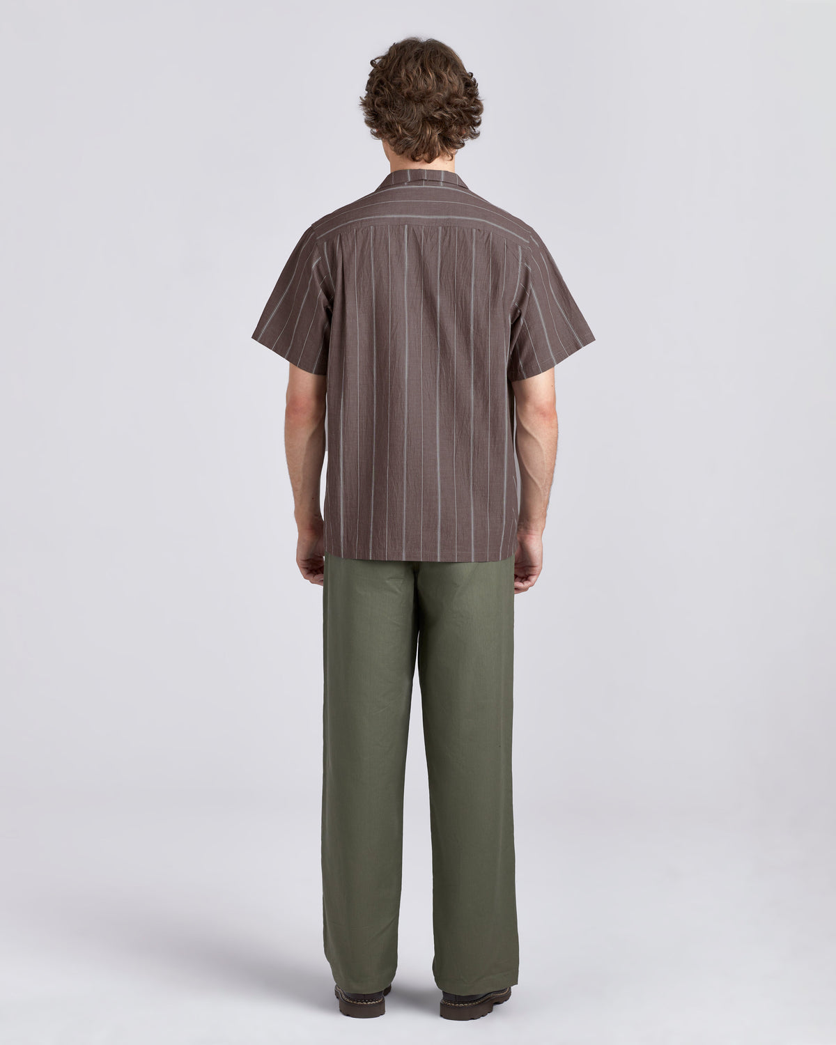A man viewed from the back wearing a brown short-sleeve button-up shirt with thin vertical white pinstripes and olive green trousers, against a light gray studio background.