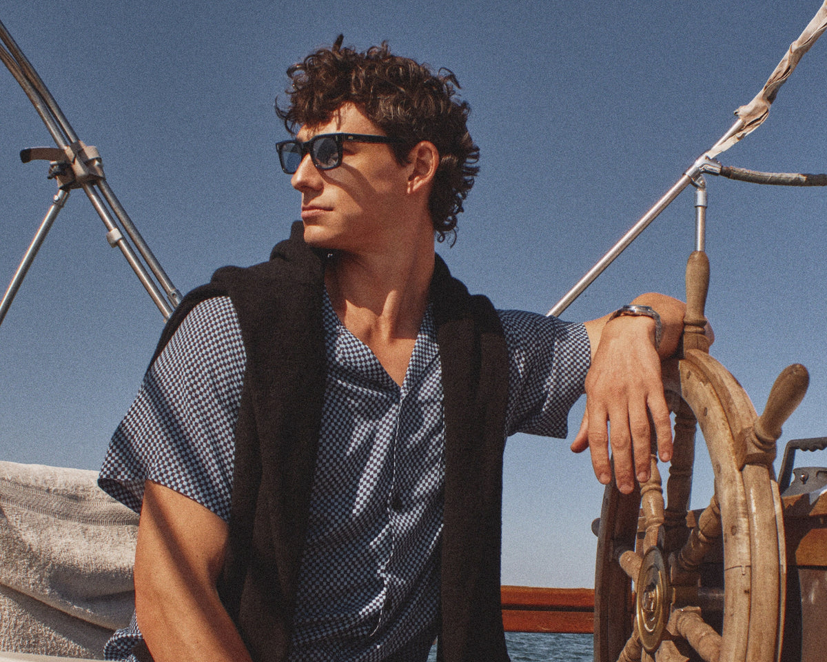 A man wearing a blue and white checkered short-sleeve shirt and matching shorts sits on a boat deck next to a wooden ship's wheel, with a black vest draped over his shoulders under a clear blue sky.