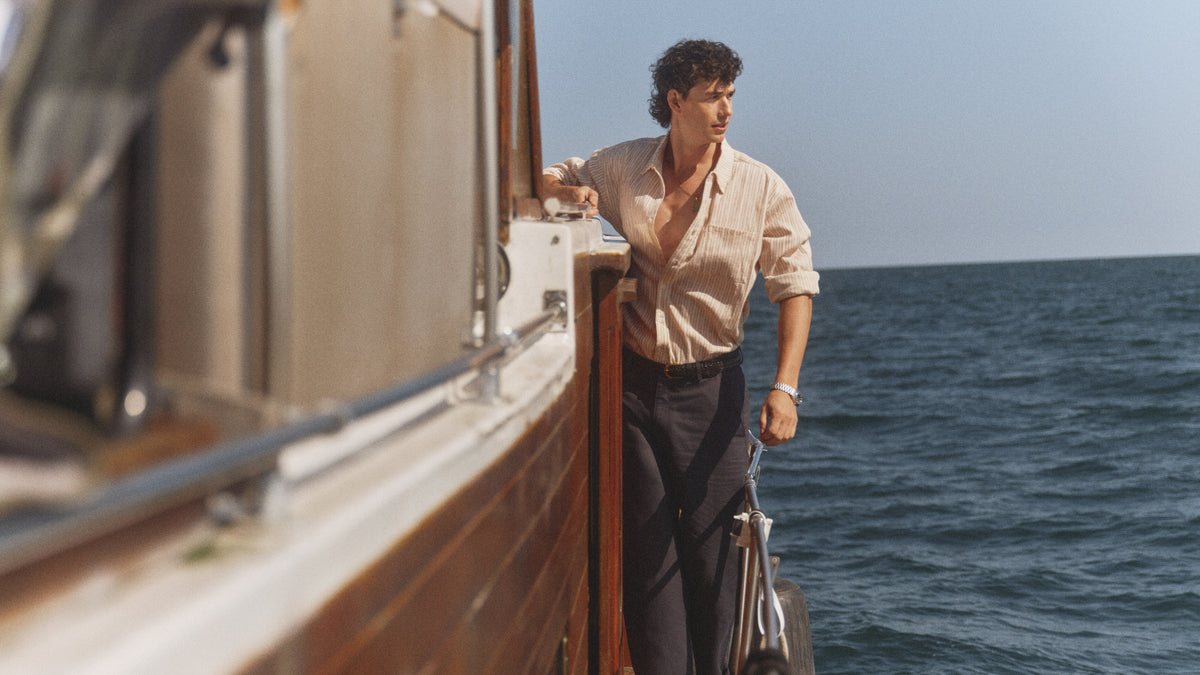 A man stands on the deck of a wooden boat on the open sea, wearing a light beige button-down shirt and dark trousers.
